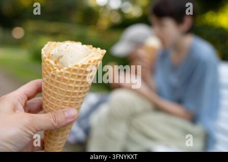 Un enfant heureux tient une corne de glace avec une boule de vanille tout en savourant une douceur dans un parc ensoleillé. Des enfants flous mangeant des sucreries en arrière-plan soulignent la modération dans l'alimentation des enfants. Banque D'Images