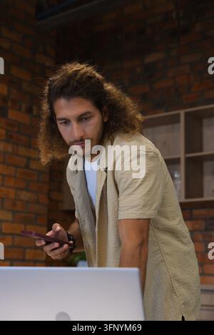 Homme d'une vingtaine d'années penché sur un ordinateur portable à un petit espace de travail, tenant un smartphone sombre, étagères en bois Banque D'Images