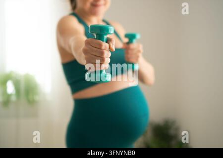 Femme enceinte tenant des haltères devant pour un léger exercice à domicile. La mère enceinte fait un entraînement doux des bras dans un espace intérieur lumineux. La future maman reste équilibrée grâce à une routine d'entraînement sûre pour débutants Banque D'Images
