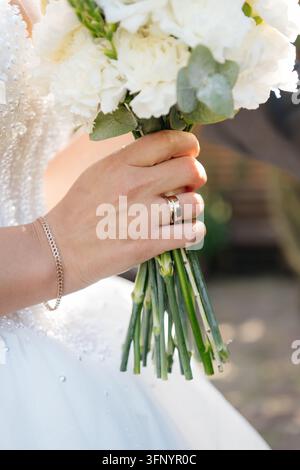 Gros plan de la main de la mariée portant une bague de fiançailles en diamant et une alliance en or, serrant les tiges vertes d'un bouquet de mariée blanc. Banque D'Images