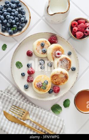 Crêpes au fromage frais avec des myrtilles fraîches, des framboises et de la menthe sur une assiette, servies avec une tasse de thé, un couteau, une fourchette et une serviette en tissu à carreaux Banque D'Images