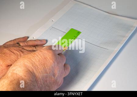 Des mains adultes tracent une ligne sur du papier quadrillé à l'aide d'un règle verte et d'un crayon jaune. Une bague en or est visible à un doigt. Banque D'Images