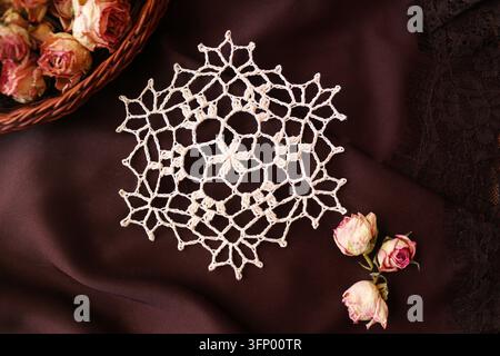 Nappette en dentelle faite à la main en forme d'étoile délicate sur un fond textile brun profond, à côté d'un panier en osier rempli de boutons de rose séchés. Banque D'Images
