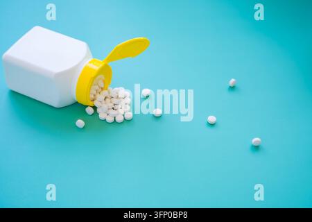 Bouteille de médecine blanche ouverte avec bouchon jaune et pilules blanches répandues sur une surface turquoise, avec des comprimés éparpillés s'étendant dans l'espace ouvert Banque D'Images