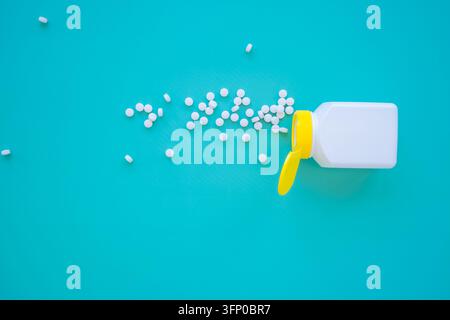 Bouteille de médicament blanche ouverte avec bouchon jaune déversant des comprimés ronds sur une surface turquoise, vue de dessus avec des pilules éparpillées et espace pour le texte Banque D'Images