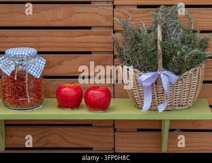 Un panier en osier contient du romarin frais attaché avec un ruban. À proximité, deux pommes rouges sont posées à côté d'un pot de pâtes sur une étagère en bois. Cette disposition met en valeur la Banque D'Images