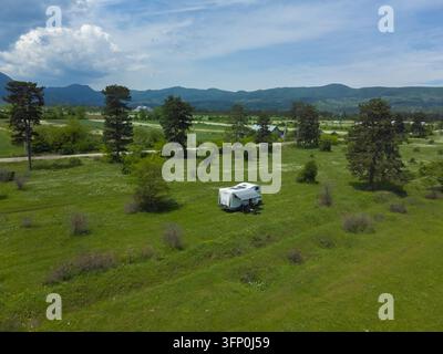 Camionnette de camping blanche profitant d'un voyage de camping en plein air, garée dans un champ vert luxuriant sous un ciel dégagé, entourée de montagnes ondulantes et d'arbres, incarnant Banque D'Images