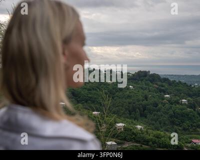 Femme blonde debout sur une colline, contemplant la vue panoramique des vastes collines vertes s'étendant jusqu'à la ville côtière et la mer sous un ciel nuageux Banque D'Images