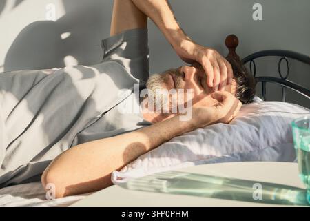 Homme allongé dans un lit souffrant d'un mal de tête, se couvrant les yeux du soleil Banque D'Images
