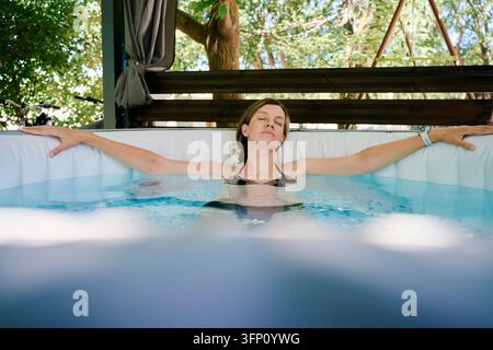 Femme se détendant dans un jacuzzi extérieur les yeux fermés. Femme reposant dans l'eau les bras étendus le long du bord de la piscine. Spa à domicile, détente estivale et bien-être Banque D'Images