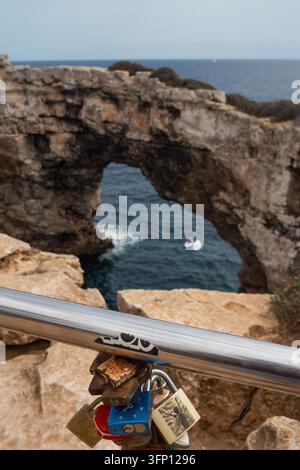 Une vue rapprochée d'une rambarde en métal ornée de nombreux cadenas d'amour, surplombée par l'arche naturelle de la mer (Es Pontas) et le bleu profond de la Méditerranée Banque D'Images