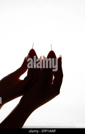 Silhouette sombre de mains féminines avec de longs ongles tenant deux bougies faites à la main en forme de fraise contre un fond blanc très lumineux avec un ba... dramatique Banque D'Images