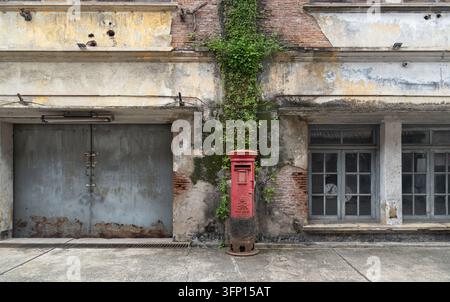 Bangkok, Thaïlande - 3 oct 2026 - Un boîte postale rouge vintage se dresse au centre devant une façade de bâtiment en briques fortement érodée, encadrée par du lierre vert rampant Banque D'Images