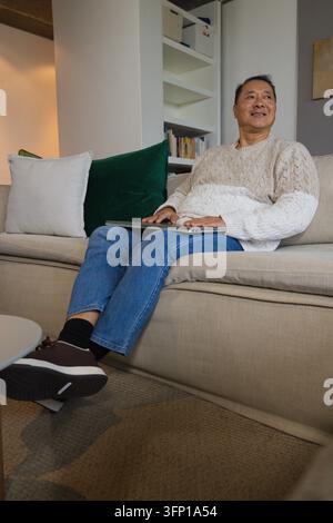 Homme chinois âgé assis sur un canapé beige dans un salon confortable utilisant un ordinateur portable portant un pull crème Banque D'Images