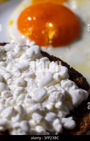 Gros plan de fromage frais sur du pain complet avec un œuf frit et un jaune d'orange coulant en arrière-plan, petit-déjeuner riche en protéines Banque D'Images
