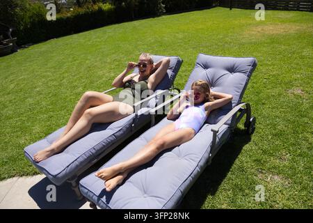 Mère et fille adolescente allongées sur des transats dans l'arrière-cour, portant des maillots de bain et des lunettes de soleil Banque D'Images