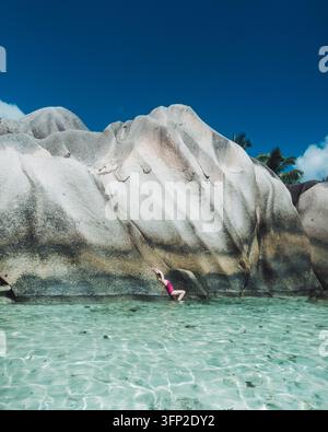 Vue panoramique d'un nageur dans une eau turquoise claire sous des rochers de granit dramatiques sur La Digue. Banque D'Images