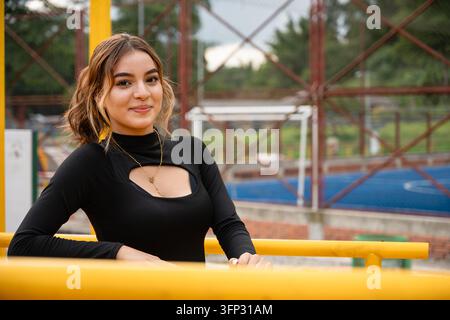 Jeune femme colombienne souriant à l'appareil, portant un haut noir avec un design découpé, appuyée sur une rambarde jaune dans un cadre de parc extérieur avec un Banque D'Images