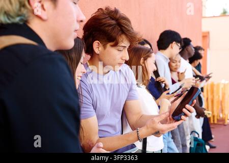 Groupe divers de jeunes étudiants debout en rangée, utilisant leurs téléphones portables et tablettes, se concentrant sur du contenu numérique Banque D'Images