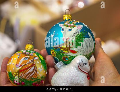 Vue rapprochée d'une main tenant des ornements de Noël en verre festifs, y compris des boules peintes en forme de chèvre et des boules blanches en forme de mouton Banque D'Images