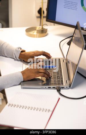 Ouvrière portant un pull à côtes tapant sur un ordinateur portable argenté à un bureau, tenant un stylo bleu près d'un carnet Banque D'Images
