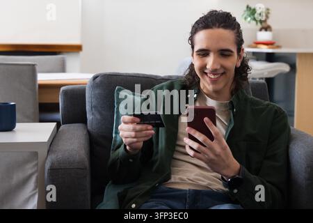 Homme afro-américain assis sur un canapé gris tenant un smartphone rouge et une carte de crédit, souriant. Téléphone, paiement, maison, canapé, minimal, moderne, bonheur Banque D'Images