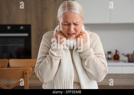 Douleur à la gorge et maladie saisonnière, femme âgée souffrant de symptômes de rhume ou de grippe à la maison avec des douleurs au cou et un inconfort dans un environnement de cuisine confortable Banque D'Images