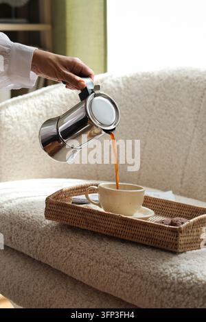 Rituel du café du matin et vie lente, personne versant un espresso frais d'une cafetière à filtre dans une tasse en céramique sur un plateau en rotin dans un cadre intérieur confortable Banque D'Images