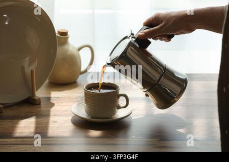Routine du café du matin et style de vie, personne versant un espresso frais d'une cafetière moka dans une tasse en céramique sur une table en bois dans un intérieur de cuisine baigné de soleil Banque D'Images