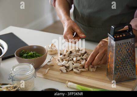 Femme mûre portant un tablier vert et coupant des champignons blancs sur une planche dans la cuisine avec un couteau d'épluchage Banque D'Images