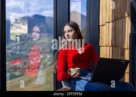 Jeune femme travaillant sur un ordinateur portable près d'une fenêtre panoramique, tenant une tasse de café tout en profitant de la lumière du soleil et d'une vue panoramique sur les montagnes dans un intérieur moderne. Banque D'Images