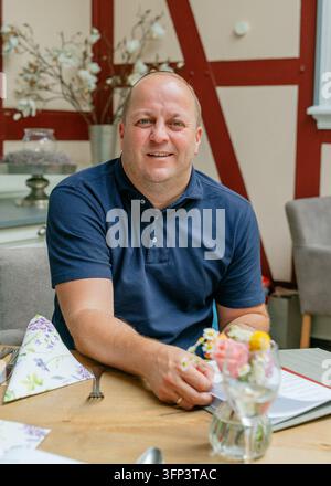 Un homme assis à une table dans un espace de repas détendu avec des décorations florales et une table en bois, souriant et regardant directement la caméra, entouré Banque D'Images