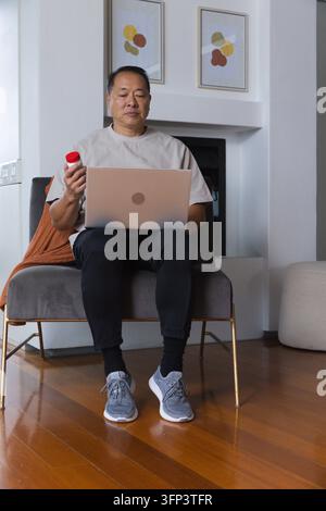 Homme asiatique âgé assis dans le salon sur une chaise grise, utilisant un ordinateur portable et tenant une bouteille à bouchon rouge Banque D'Images