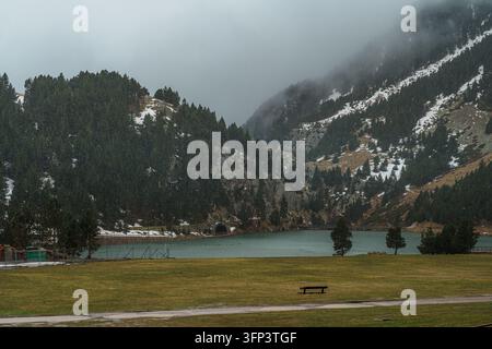Un banc solitaire se dresse sur une prairie vide devant un lac de montagne vert brumeux et des pentes boisées de pins striées de neige à Vall de Nuria, Pyrénées, Espagne Banque D'Images