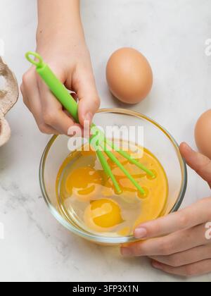 Enfant fouettant des œufs cassés dans un bol en verre transparent avec un fouet en plastique vert, gros plan des mains sur la table de la cuisine Banque D'Images