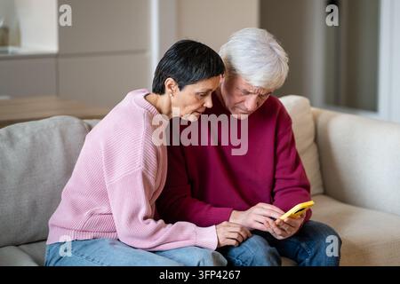Couple de personnes âgées positives intéressées assis ensemble sur un canapé en parcourant des médias en ligne sur un smartphone. Banque D'Images