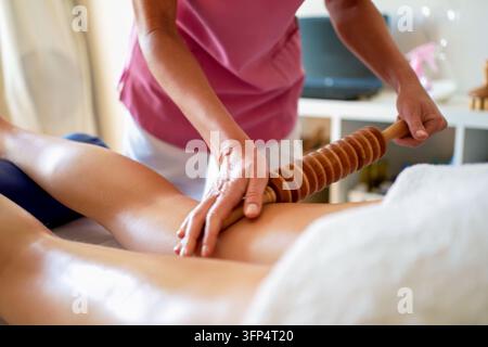 Thérapeute utilisant un rouleau en bois sur le mollet du client lors d'une séance de massage sereine dans une salle de traitement chaude avec une pression manuelle concentrée et un bien-être calme Banque D'Images