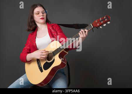 Jeune guitariste confiante qui strumming guitare acoustique et regardant vers le bas les cordes avec concentration. Musicien élégant de femme portant des lunettes, d Banque D'Images