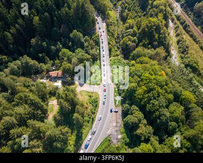 La campagne boisée de Roumanie entoure une route sinueuse animée par des voitures dans les deux sens. Une vue élevée montre la canopée forestière, les lignes utilitaires du bord de route et sm Banque D'Images