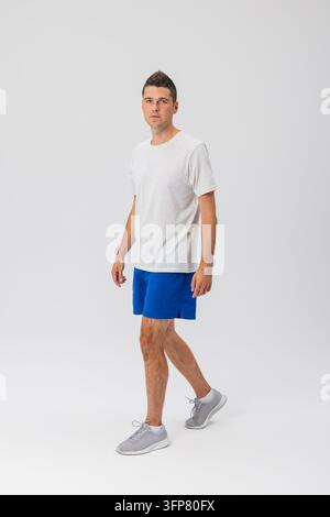 Portrait en corps entier d'un homme athlétique mince marchant sur le côté contre un fond blanc isolé du studio, portant un t-shirt gris clair et un athle bleu Banque D'Images