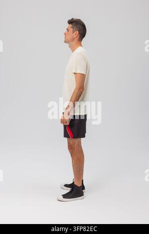 Vue de côté en pleine longueur d'un jeune homme de profil, portant un t-shirt crème, un short de sport noir avec des bordures rouges et des baskets sur un fond gris Banque D'Images