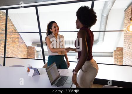 Diverses collègues femmes se serrant la main à travers une table de réunion avec un ordinateur portable ouvert, une tablette, un verre, des papiers Banque D'Images