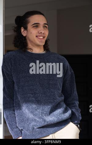 Homme afro-américain début de la vingtaine appuyé contre un cadre de porte blanc en souriant, portant un pull bleu marine Banque D'Images