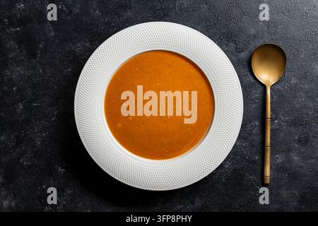 Soupe à la citrouille flat lay sur fond sombre avec cuillère dorée. Vue de dessus de la soupe crémeuse orange dans une assiette blanche texturée. Minimaliste saine citrouille ou tomate Banque D'Images