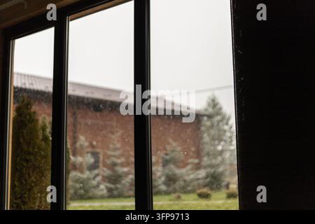 Des gouttes de pluie recouvrent une grande vitre aux cadres sombres. Un bâtiment en briques flou, des arbres à feuilles persistantes et un ciel nuageux apparaissent à l'extérieur du verre Banque D'Images