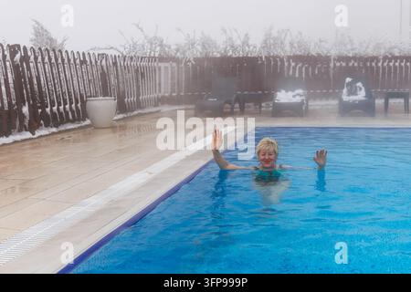 • Femme mûre heureuse portant des lunettes agitant les mains tout en se détendant dans une piscine thermale extérieure chauffée d'un complexe hôtelier luxueux en montagne. Banque D'Images