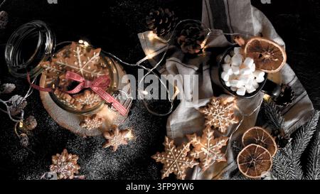 Des biscuits en forme de flocon de neige décorés reposent sur une surface rustique sombre à côté d'un bocal de bonbons, d'une tasse de guimauves, de tranches de citron séchées et d'une lumière féerique scintillante Banque D'Images