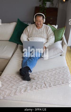 Homme asiatique âgé portant un pull en câble crème et des écouteurs blancs utilisant un ordinateur portable argenté à la maison Banque D'Images