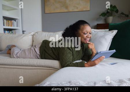 Sénior africaine allongée sur un canapé d'angle à la maison portant un pull olive tenant une tablette Banque D'Images