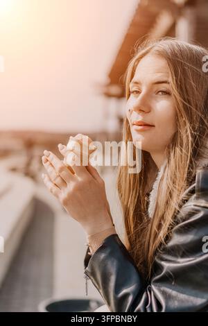 Femme avec glace au coucher du soleil, blonde, jeune femme en veste en cuir tenant un cône à dessert tout en étant assise à l'extérieur à l'heure dorée Banque D'Images